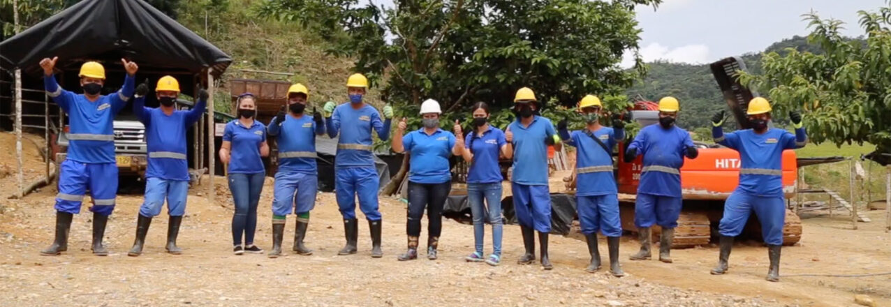 Group of men and women gold miners in blue uniforms and yellow helmets standing in front of mining equipment in a mining area on a rocky shore in a rainforest in Antioquía, Colombia.