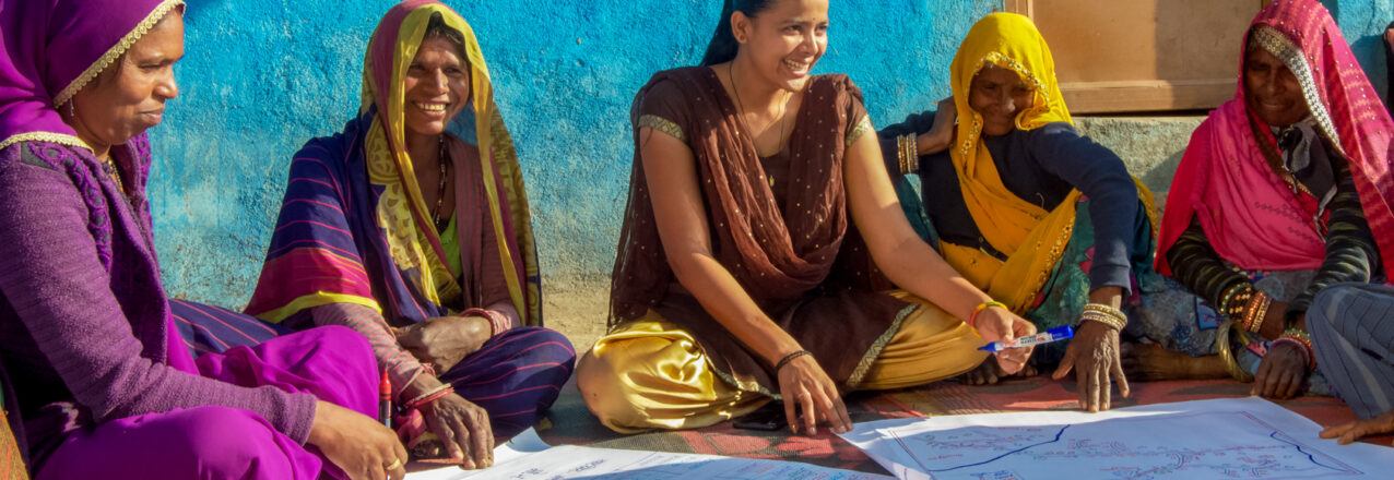 Five women sit on a mat and study several large documents that are laid out in front of them.